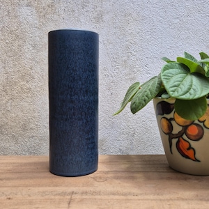 Könnte beinhalten: Eine hohe, zylindrische Vase in einer gesprenkelten marineblauen Glasur. Die Vase steht auf einer Holzoberfläche, im Hintergrund ist ein dekorativer Blumentopf zu sehen. Die Vase ist ein dekorativer Artikel für das Zuhause.