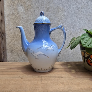 May include: A porcelain teapot with a light blue gradient, featuring a gold trim and a lid with a decorative finial. The teapot is adorned with flying seagull illustrations. The handle and spout are elegantly designed.