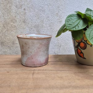 May include: A handmade ceramic planter with a light brown and pink speckled finish. The planter has a slightly flared shape and sits on a wooden surface. A second planter with green leaves is partially visible.