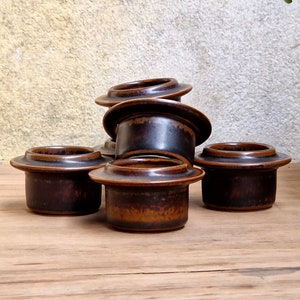 Może przedstawiać: Sześć brązowych ceramicznych stojaków na jajka ułożonych na drewnianej powierzchni. Stojaki na jajka mają ciemnobrązową glazurę z jaśniejszym brązowym paskiem wokół górnej części.