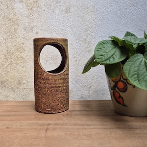 Könnte beinhalten: Eine zylindrische, braune Keramikvase mit einem großen kreisförmigen Ausschnitt. Die Vase hat eine strukturierte Oberfläche und steht auf einer Holzoberfläche. Eine Topfpflanze mit grünen Blättern befindet sich im Hintergrund.