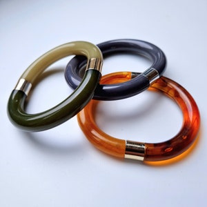 Puede incluir: Tres pulseras de plástico en diferentes colores: verde oliva, morado y ámbar. Las pulseras están apiladas una encima de la otra.