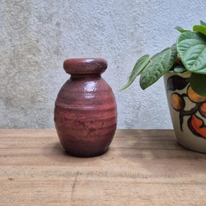 Puede incluir: Un pequeño jarrón artesanal de cerámica de color marrón rojizo, con un cuerpo redondeado y un cuello estrecho. El jarrón tiene una superficie texturizada y parece estar esmaltado. El jarrón está sobre una superficie de madera.