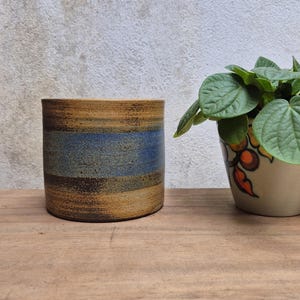 May include: A cylindrical ceramic planter with a textured surface. The planter features horizontal bands of brown, blue, and tan. A small potted plant with green leaves sits to the right of the planter on a wooden surface.