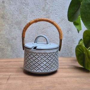 Puede incluir: Azucarero de cerámica azul claro con tapa y asa trenzada. El cuenco tiene un patrón repetitivo y está sobre una superficie de madera. El asa es de ratán trenzado. Es un artículo de menaje.