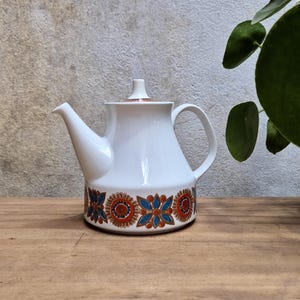 May include: White ceramic teapot with a lid and handle. The base features a repeating floral pattern in orange, blue, and brown. The teapot is set on a wooden surface with a neutral background and a plant in the background.