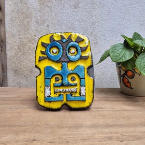 Peut inclure: Une pièce d'art en céramique jaune et bleue avec un motif de visage stylisé. La pièce présente un fond jaune avec des traits du visage bleus et des accents blancs. L'œuvre est placée sur une surface en bois.