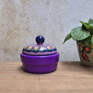 May include: A purple ceramic jar with a lid. The lid is decorated with a floral pattern in yellow, pink, and blue, and topped with a blue knob. The jar is on a wooden surface.