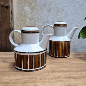 Könnte beinhalten: Zwei weiße Keramikkannen mit braunen und orangefarbenen Streifenmustern. Die Kannen haben einen Ausguss und einen Henkel.