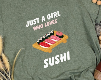 May include: Green t-shirt with the text "JUST A GIRL WHO LOVES SUSHI" and a sushi graphic. The shirt is displayed with brown shoes, wooden beads, and a decorative woven basket on a plaid blanket.