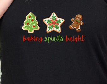 May include: Black t-shirt featuring a Christmas-themed design. The design includes a gingerbread tree, star, and man, with the phrase "baking spirits bright" in red script. The shirt is a crew neck.