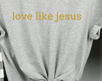 May include: A gray t-shirt with the text "love like jesus" in gold lettering. The shirt is knotted at the waist and hangs on a wooden hanger. A black and white plaid shirt and blue jeans are also on hangers.