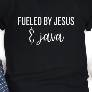 May include: Black t-shirt with the white text "Fueled by Jesus & Java". The text is in a bold, sans-serif font, with the word "Java" in a cursive script. The shirt is folded, with a glimpse of blue patterned fabric and a black shoe visible.