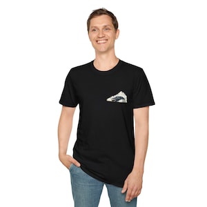 Peut inclure: T-shirt noir avec un petit motif sur la poitrine gauche. Le motif représente une voiture sombre entourée de fumée blanche. La personne sur l'image porte un jean bleu.