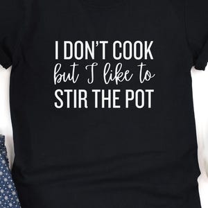 May include: Black t-shirt with the white text "I DON'T COOK but I like to STIR THE POT". The shirt is a crew neck style and appears to be made of a soft material. The shirt is displayed on a white background.