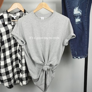 May include: A grey t-shirt with the text "it's a good day to code" is knotted at the waist. A black and white checked shirt and distressed blue jeans hang on a clothes rail. A brown hat is visible.