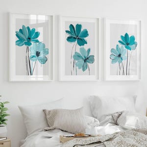 May include: A set of three framed wall art prints featuring teal blue flowers with dark stems on a light grey background. Each vertical rectangular frame contains a botanical illustration of two or three blossoms with visible brushstroke textures.