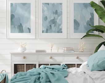 May include: Three framed vertical wall art prints featuring abstract brushstrokes in shades of light blue and white. The artwork is displayed above a white wooden console table with drawers and woven baskets, alongside a large green potted plant.