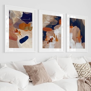 May include: Three abstract paintings in white frames with a beige, brown, and blue colour scheme. The paintings are hung on a white wall above a white sofa with cushions.