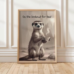 May include: A framed print showing a meerkat holding a teacup, with steam rising. The sepia-toned image includes the text "On the lookout for tea!". The artwork is set against a neutral background and framed in light wood, suitable for a home or office.