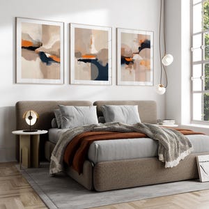 May include: Three framed abstract art prints with beige, orange, and navy blue color blocks hang above a bed. The bed has gray bedding, a brown blanket, and a patterned throw. A small side table with a lamp sits next to the bed.