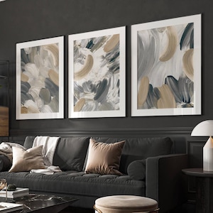 Könnte beinhalten: Drei gerahmte abstrakte Kunstdrucke. Jeder Druck zeigt Pinselstriche in Grau-, Beige- und Weißtönen. Das Kunstwerk wird über einem dunkelgrauen Sofa mit dekorativen Kissen ausgestellt. Die Rahmen haben einen weißen Rand.