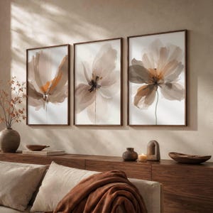 May include: A set of three vertical wall art prints featuring abstract floral illustrations in shades of beige, cream, and soft orange. Each rectangular piece is enclosed in a thin dark brown frame, displaying translucent, layered flower petals.