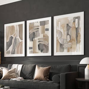 Könnte beinhalten: Drei abstrakte Kunstdrucke in weißen Rahmen mit einem Farbschema in Braun, Grau und Weiß. Die Drucke hängen an einer dunkelgrauen Wand über einem grauen Sofa.
