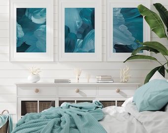 May include: Three framed vertical wall art prints featuring abstract brushstrokes in shades of teal, navy, and white. The artwork is displayed in white frames above a white wooden console table with drawers and woven baskets, near a green plant.