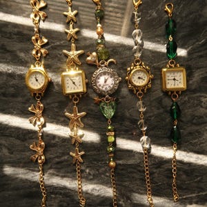 Puede incluir: Cinco relojes con colgantes dorados con varios elementos decorativos. Los relojes tienen esferas redondas o cuadradas con números romanos. Los adornos incluyen lazos, estrellas de mar, cuentas y cadenas. Los relojes se exhiben sobre una superficie oscura y texturizada.
