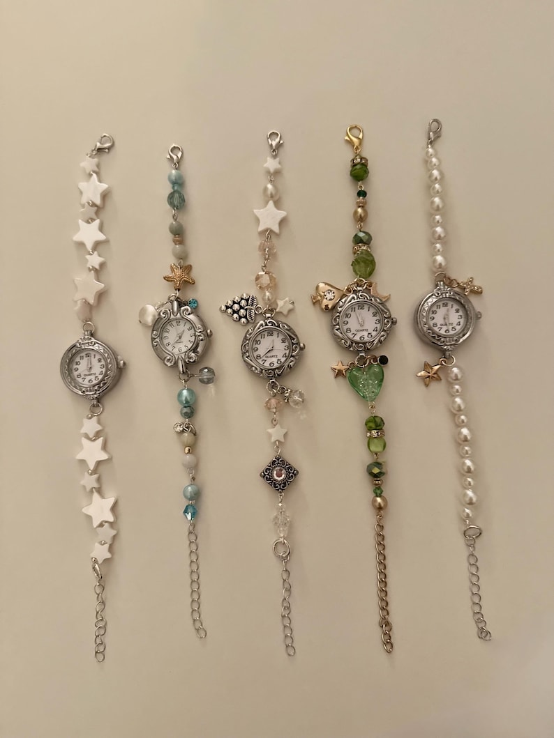 May include: Five decorative wristwatches with silver-toned cases and white watch faces. Each watch features a unique beaded or chain strap with charms, including stars, pearls, and various colored beads. The watches are displayed against a neutral background.