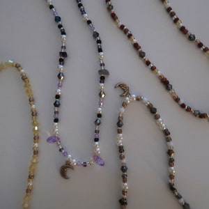 May include: Four beaded necklaces with crescent moon charms. The necklaces are made with various colors of beads, including white, brown, black, purple, and gold.