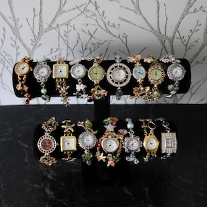 Può includere: Una collezione di diciassette orologi da polso decorativi esposti su un supporto nero a due livelli. Gli orologi presentano varie finiture metalliche, toni oro e argento, con diverse forme di quadrante, perline colorate e ciondoli.