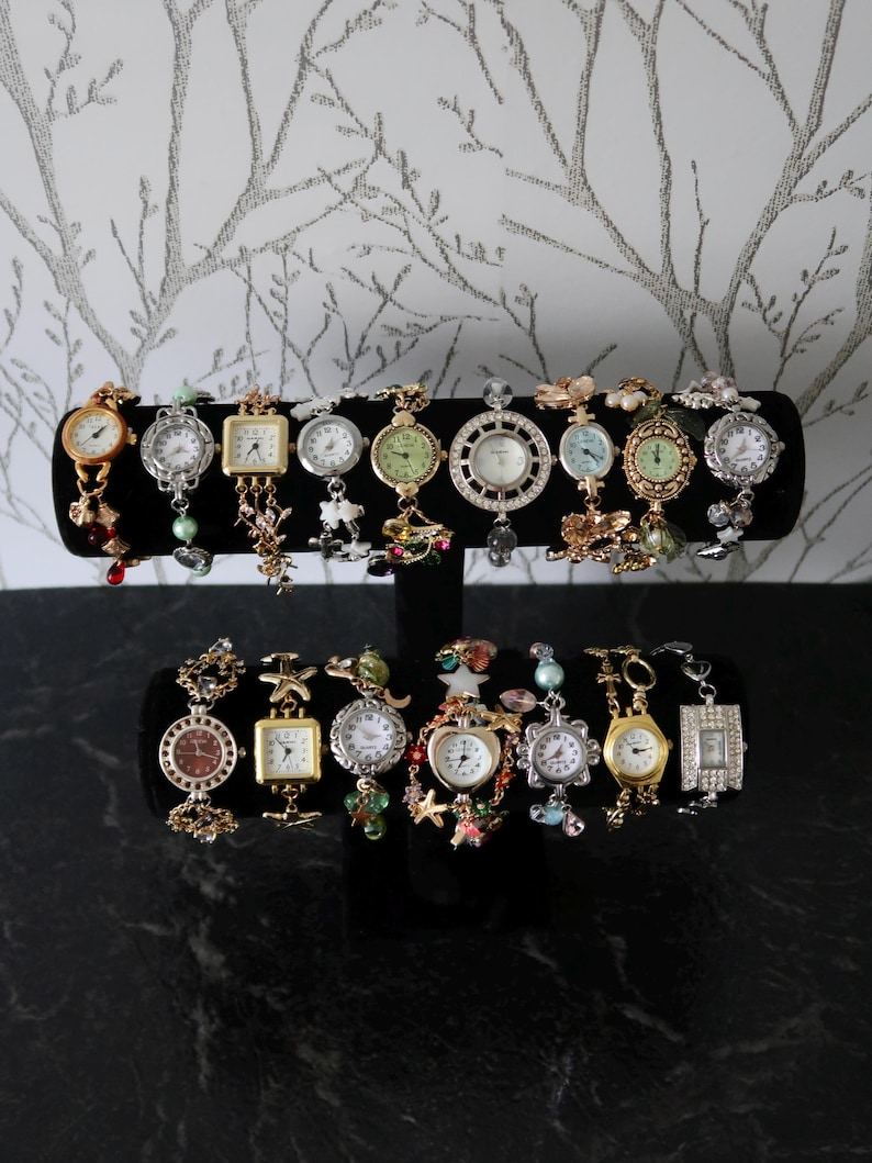 Puede incluir: Una colecci&oacute;n de diecisiete relojes de pulsera decorativos expuestos en un soporte negro de dos niveles. Los relojes presentan diversos acabados met&aacute;licos, tonos dorados y plateados, con formas de esfera variadas, cuentas y abalorios.