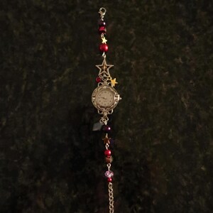 May include: A silver-toned watch with a white face, adorned with red and purple beads, and star charms. The watch has a chain and a decorative star at the top. The watch is a unique accessory.