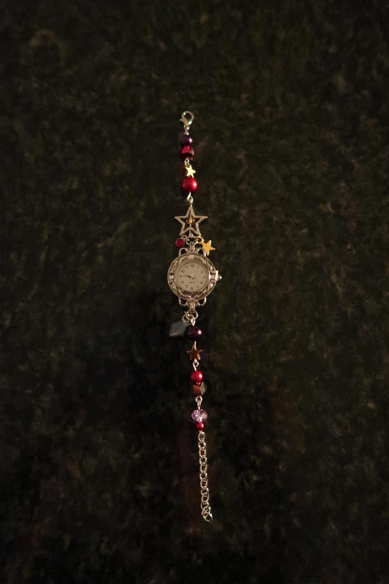 May include: A silver-toned watch with a white face, adorned with red and purple beads, and star charms. The watch has a chain and a decorative star at the top. The watch is a unique accessory.