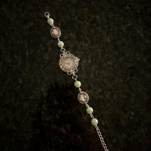 May include: A silver watch with a white face and black numbers. The watch is attached to a silver chain with green beads.