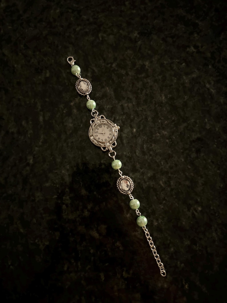 May include: A silver watch with a white face and black numbers. The watch is attached to a silver chain with green beads.