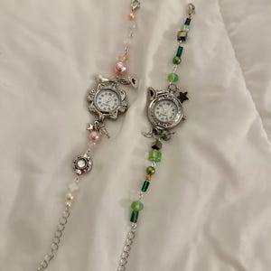 Puede incluir: Dos pulseras de plata con abalorios de diferentes colores. Una pulsera tiene cuentas rosas y blancas, la otra tiene cuentas verdes y doradas. Ambas pulseras tienen una pequeña esfera de reloj de plata con números romanos.