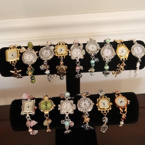 Op de afbeelding: Een display van 12 zilveren en gouden armbandjes met kleine ronde horloge-wijzerplaten. De armbandjes zijn versierd met kralen en bedels, waaronder sterren, bloemen en andere decoratieve elementen.