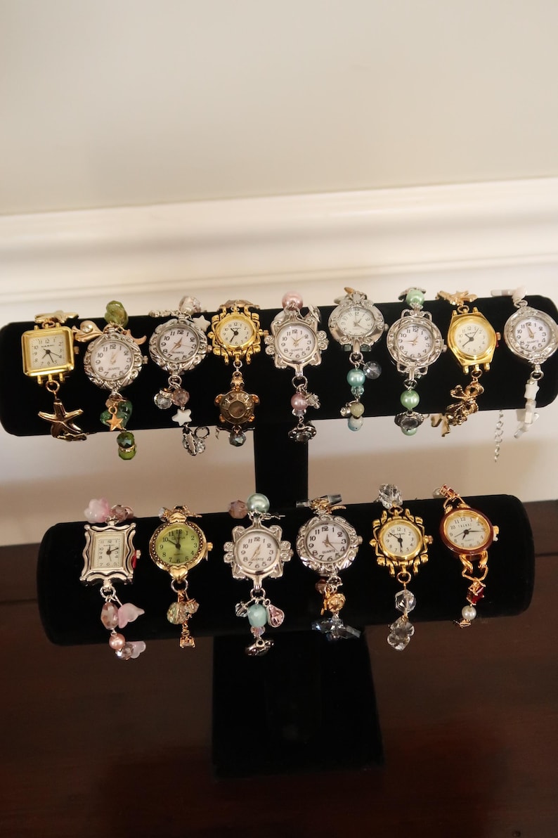Op de afbeelding: Een display van 12 zilveren en gouden armbandjes met kleine ronde horloge-wijzerplaten. De armbandjes zijn versierd met kralen en bedels, waaronder sterren, bloemen en andere decoratieve elementen.
