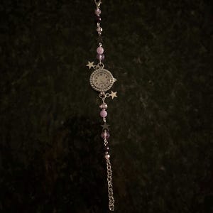 May include: A silver-toned watch with a white face and black numerals, adorned with purple and black beads, and star charms. The watch band features a chain and a clasp. The watch is set against a dark background.