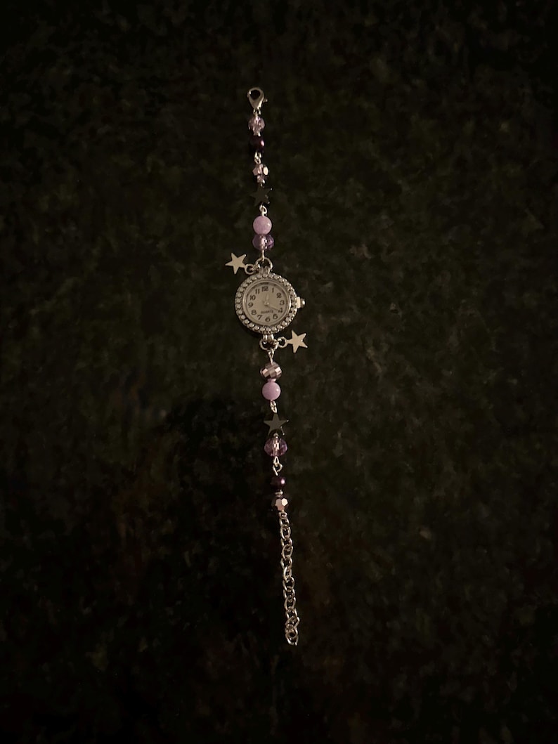 May include: A silver-toned watch with a white face and black numerals, adorned with purple and black beads, and star charms. The watch band features a chain and a clasp. The watch is set against a dark background.