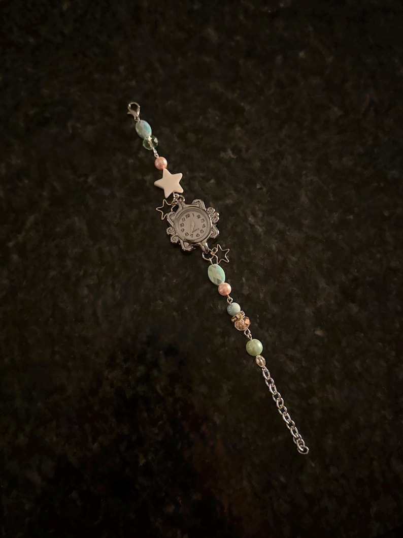 May include: A silver-toned watch with a white face and black numerals, adorned with a beaded bracelet. The bracelet features turquoise, pink, and gold-toned beads, along with star-shaped charms. A delicate chain completes the design.