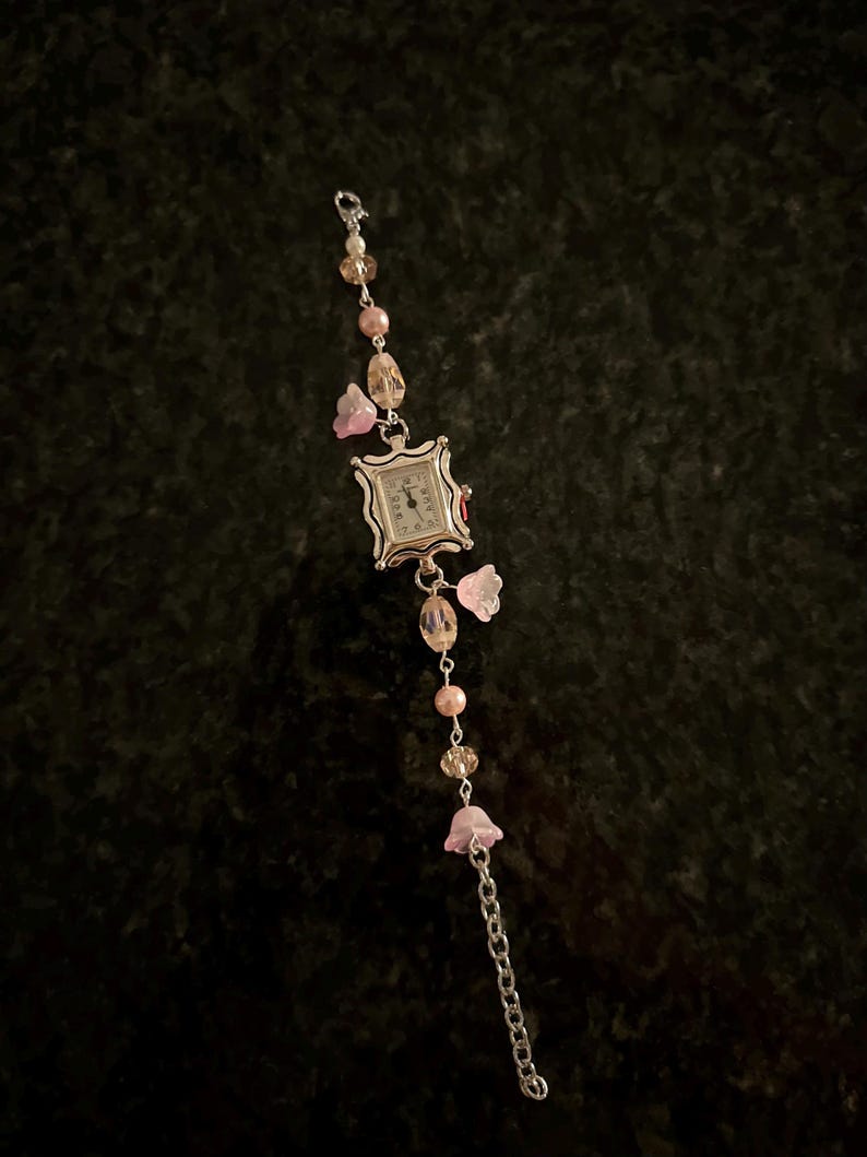 May include: A silver watch with a square face and a beaded chain strap. The beads are pink, white, and clear. The watch has a floral design with pink flowers.