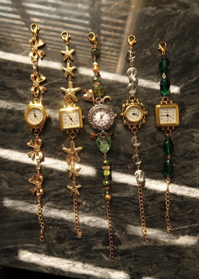 May include: Five gold-toned charm watches with various decorative elements. The watches have round or square faces with Roman numeral hour markers. Embellishments include bows, starfish, beads, and chains. The watches are displayed on a dark, textured surface.