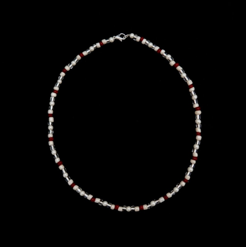 May include: A silver and white beaded necklace with red accents. The necklace is made of small, round beads and has a simple clasp.