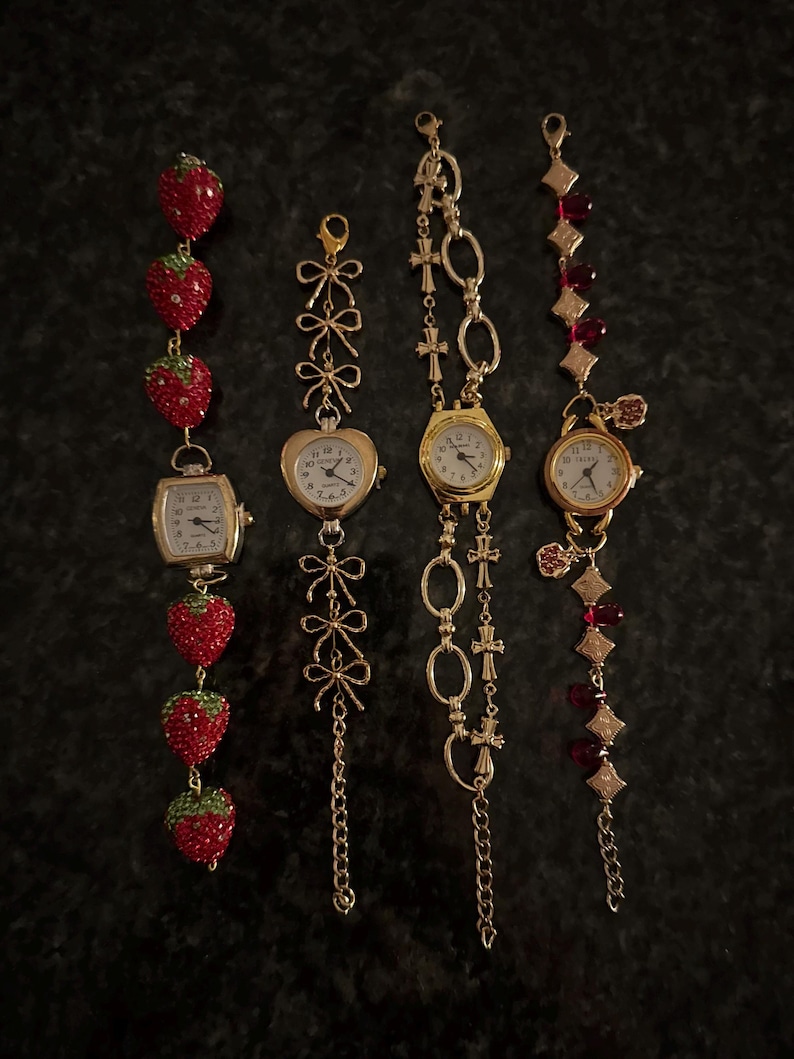 May include: Four decorative wristwatches with gold-toned metal chains. One watch features a strawberry design, another has a heart-shaped face, and two others have round faces. The watch faces have white backgrounds and black numbers.