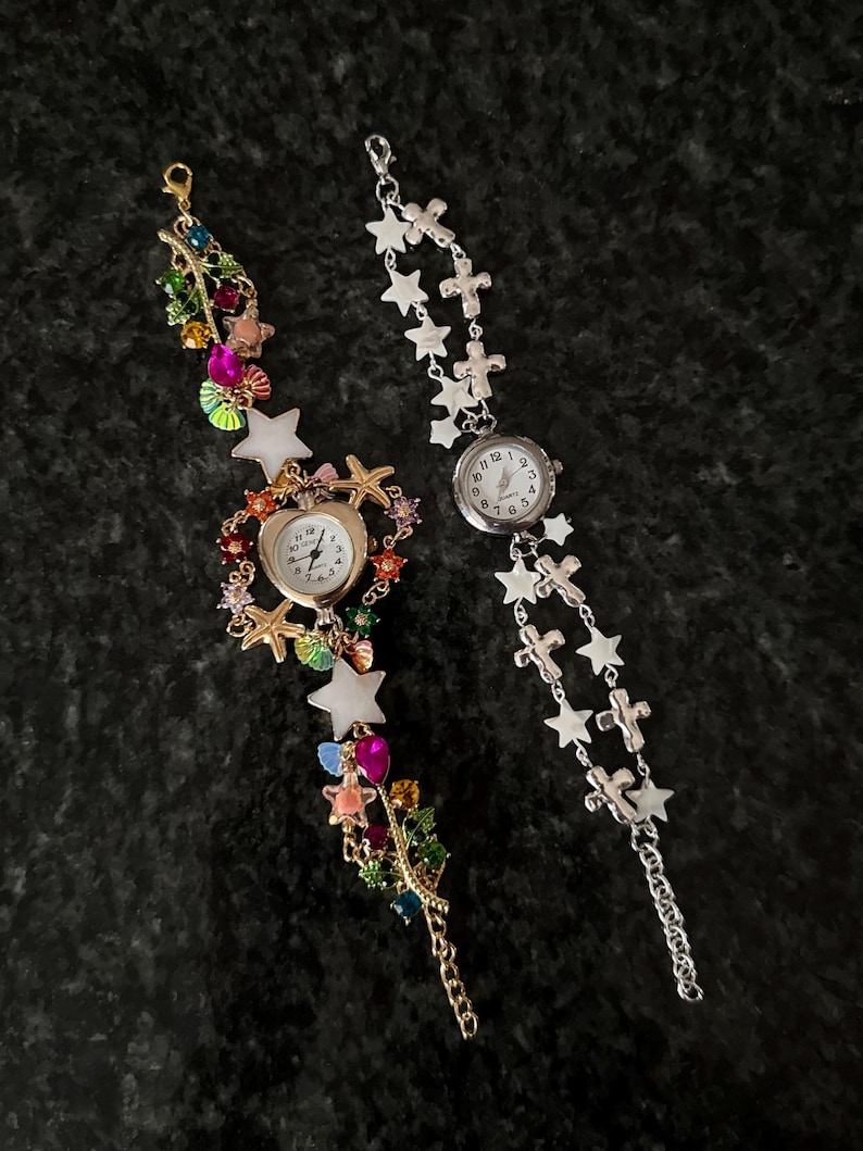 May include: Two wristwatches with decorative chains. The left watch features a heart-shaped face surrounded by colorful star, shell, and gemstone charms. The right watch has a round face with a silver-tone chain featuring alternating star and cross charms.