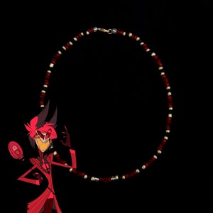May include: A red and black beaded necklace with a gold clasp. The necklace is circular in shape and is hanging against a black background.